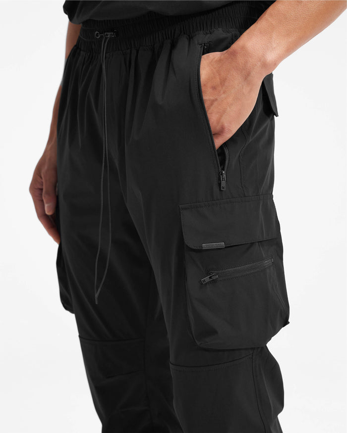 Man wearing black cargo pants on a plain white background | 247 Pant in Black | Nylon pants with cargo pockets and adjustable features | 247 Pants | Represent Clo