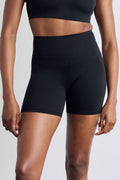 A close-up of a model wearing black high-waisted athletic shorts and a matching black sports bra against a light gray studio background.