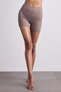 A model's lower body wearing high-waisted, seamless athletic shorts in a muted taupe-brown color, standing barefoot on a gray yoga mat against a light gray studio background.