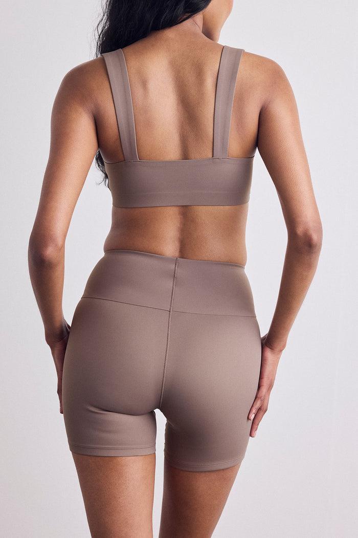 Rear view of a woman wearing a matching taupe-brown activewear set consisting of a sports bra and high-waisted biker shorts, set against a plain light gray background.