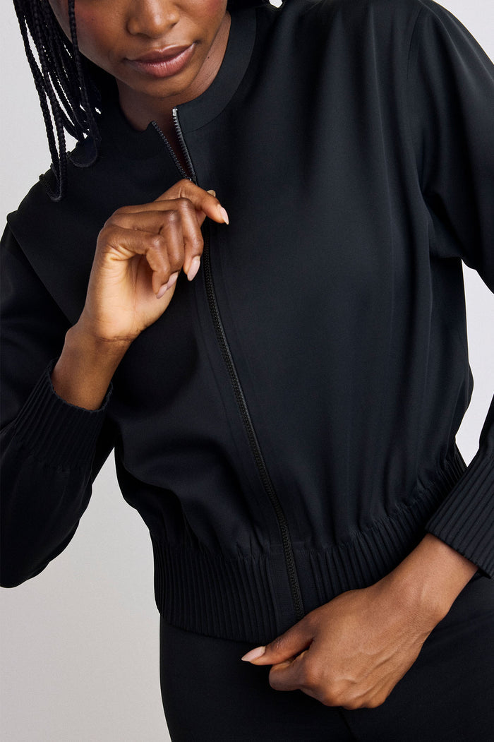 A close-up of a model wearing a black zip-up bomber jacket, showing the collar, cuffs, and hem, with one hand adjusting the zipper.