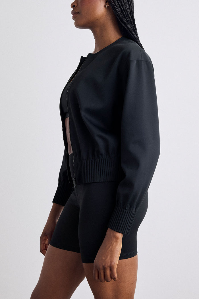 A model shown from the side wearing a black bomber jacket with ribbed cuffs and hem, paired with black fitted shorts, against a light gray studio background.