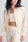 A woman wearing a matching cream-colored activewear set, featuring a cropped zip-up bomber jacket, a sports bra, and high-waisted leggings, set against a plain light gray background.