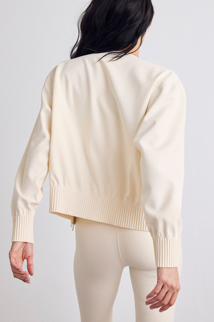 A model shown from the back wearing a cream-colored bomber with ribbed cuffs and hem, paired with matching cream leggings, against a light gray studio background.
