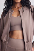 A close-up of a model wearing a matching taupe activewear set, featuring a ribbed zip-up bomber jacket, a scoop-neck sports bra, and high-waisted leggings.