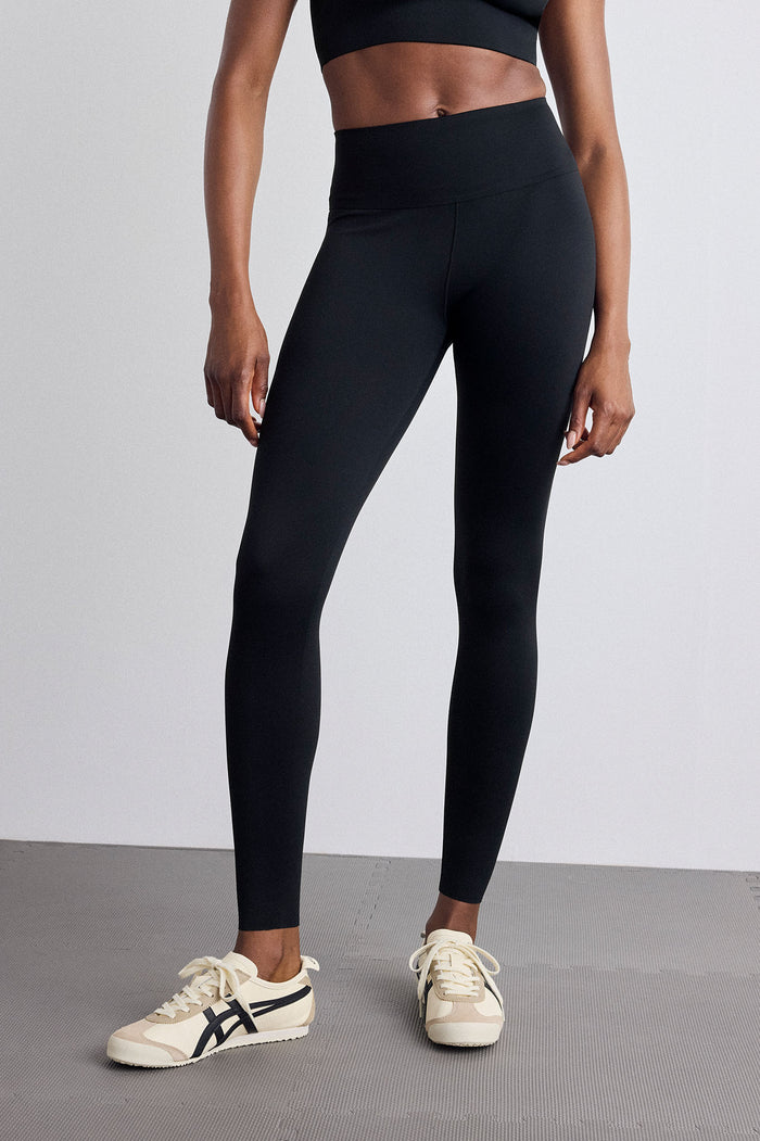 A model wearing black high-waisted athletic leggings and cream sneakers with black stripes, standing against a light gray studio background.