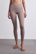 A model wearing high-waisted, full-length leggings in a muted taupe-brown color, standing barefoot on a gray floor against a light gray wall.