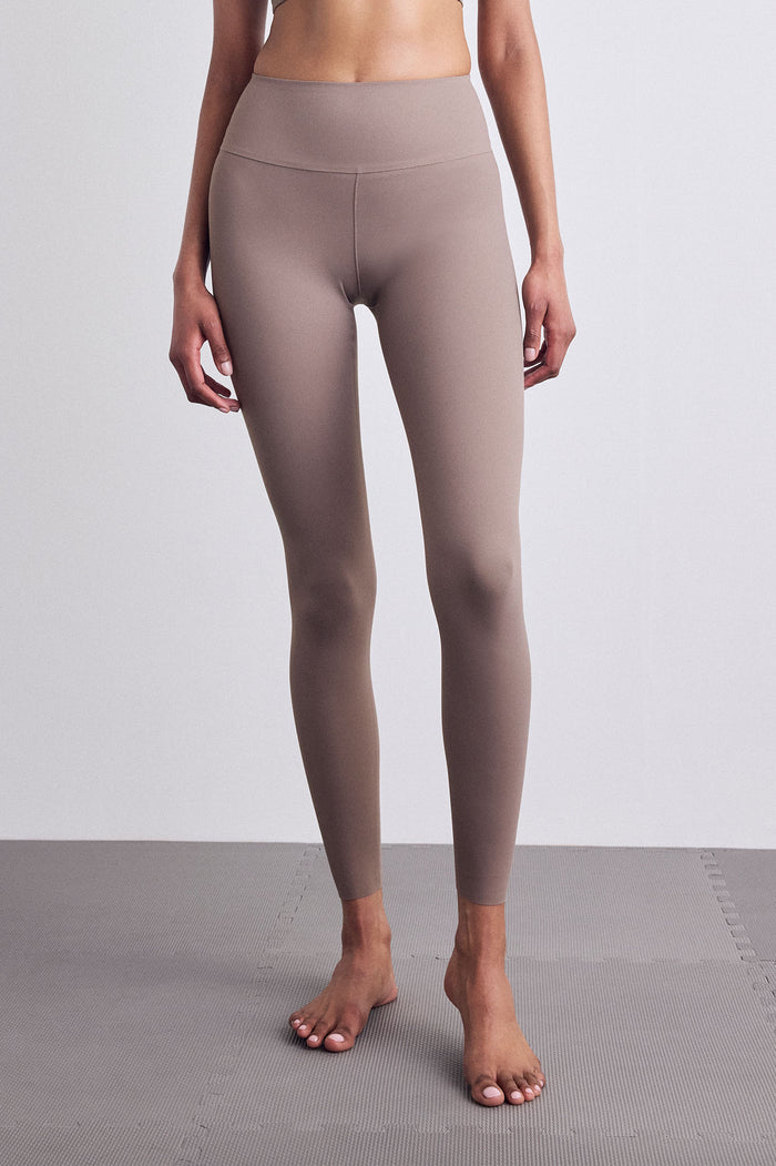 A model wearing high-waisted, full-length leggings in a muted taupe-brown color, standing barefoot on a gray floor against a light gray wall.