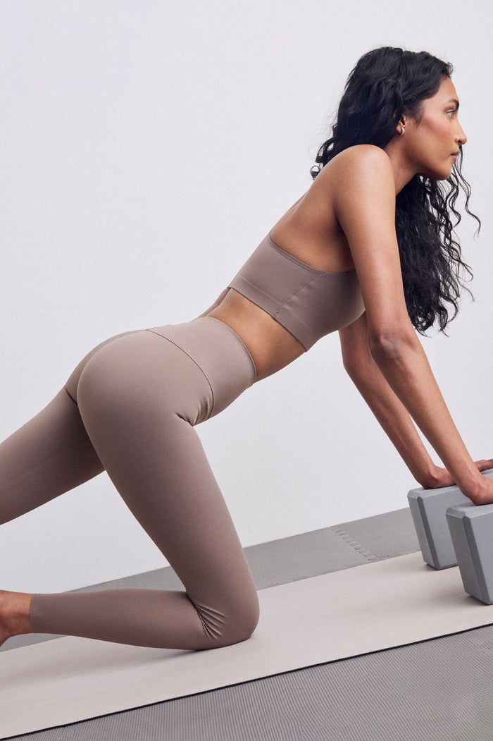 A woman in a matching taupe activewear set, consisting of a sports bra and high-waisted leggings, is in a side-lying push-up position on a light gray exercise mat with gray yoga props.