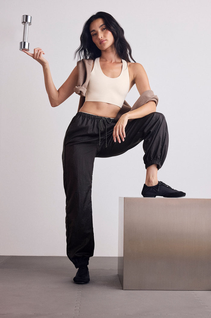 A woman poses in a studio wearing a beige sports bra, black jogger pants, and black sneakers, holding a silver dumbbell.