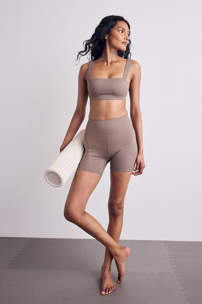 A woman modeling a matching taupe activewear set consisting of a sports bra and high-waisted shorts, holding a white textured foam roller against a neutral studio backdrop.