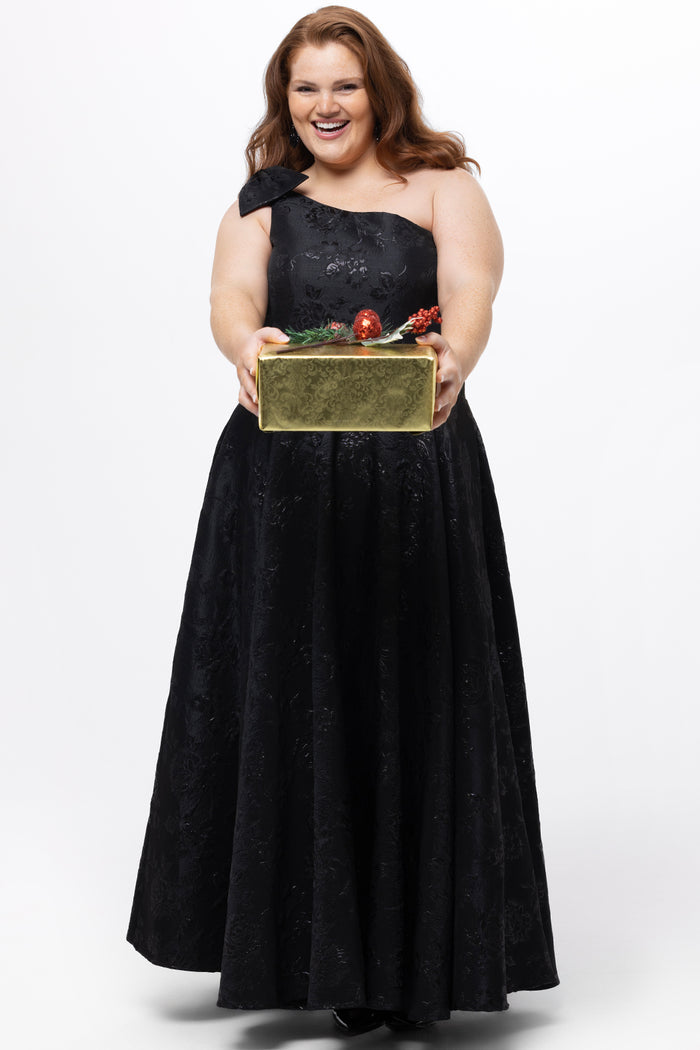 Celebrations by Sydney's Closet Style CE2614 in Black

Plus size maxi length formal dress. One-shoulder neckline with detachable bow at high point of shoulder, natural waistline and A-line skirt with pockets. 

Black floral brocade fabric. 

Shown with model holding a Christmas gift.