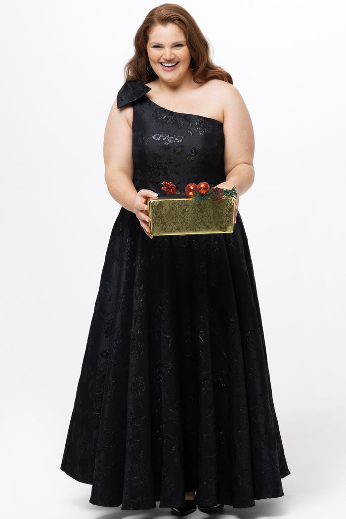 Celebrations by Sydney's Closet Style CE2614 in Black

Plus size maxi length formal dress. One-shoulder neckline with detachable bow at high point of shoulder, natural waistline and A-line skirt with pockets. 

Black floral brocade fabric. 

Shown with model holding a Christmas gift.