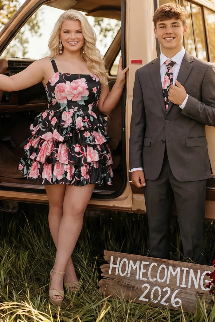 Young couple outdoors beside a vintage tan vehicle. The young woman wears a black and pink sleeveless mini dress with a pink floral print, square neckline, and tiered ruffle skirt, paired with nude platform ankle-strap heels and statement earrings. Young man wears a charcoal suit with a white dress shirt and a floral tie that matches the dress. Rustic wooden sign reading “Homecoming 2026” sits in the grass in front of them, a coordinated homecoming portrait. Dress is Sydney's Closet SC8174