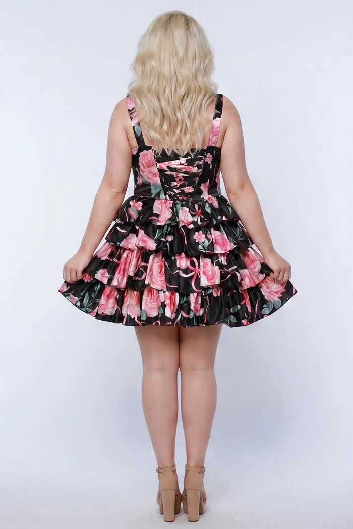 Plus-size model wearing a black and pink floral sleeveless mini dress with layered ruffle skirt. The dress features a square neckline, fitted bodice, and tiered ruffles at the waist and hem. She is styled with long wavy blonde hair, lace-up back with modesty panel, and nude platform ankle-strap heels, posing against a studio background. Back view. 

Sydney's Closet SC8174