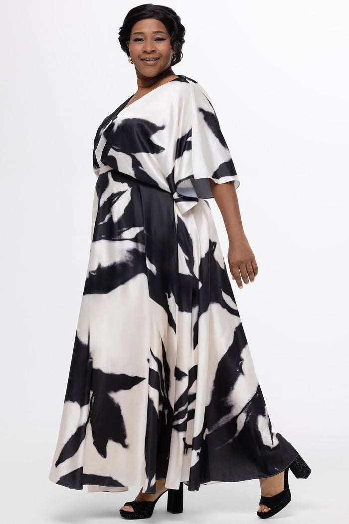 Celebrations by Sydney's Closet Style CE2620 in black/white geometric print. 

Plus-size model wearing a flowing black-and-ivory abstract print maxi dress with a V-neckline, draped bodice, elbow-length sleeves, and a softly flared skirt, photographed against a white background.