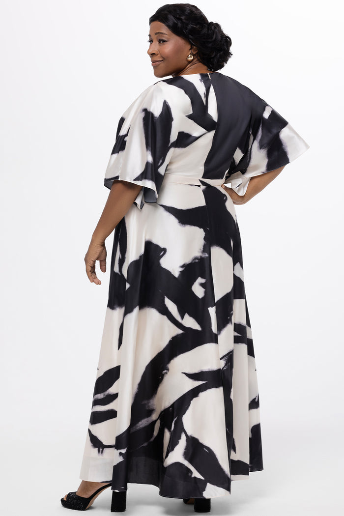Celebrations by Sydney's Closet Style CE2620 in black/white geometric print. 

Plus-size model wearing a flowing black-and-ivory abstract print maxi dress with a V-neckline, draped bodice, elbow-length sleeves, and a softly flared skirt, photographed against a white background.