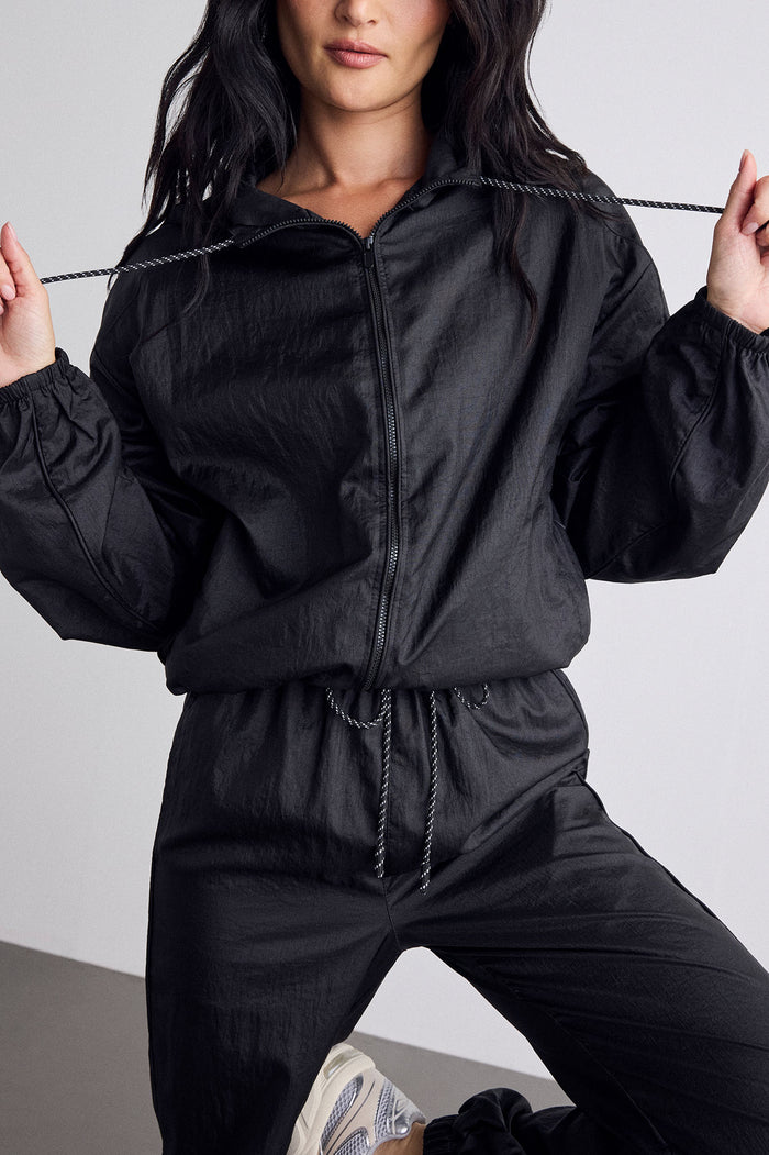 A model wearing a black zip-up windbreaker jacket and matching black drawstring sweatpants, holding the jacket's drawstrings outwards against a light gray studio background.