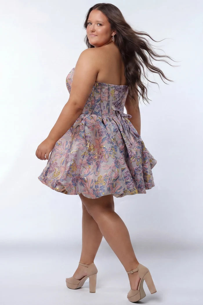 Plus-size model wearing a strapless a-line mini dress in purple floral brocade with metallic gold accents. The dress features a structured corset-style bodice with a strapless sweetheart neckline and a voluminous bubble hem skirt. She is styled with long wavy dark brown hair, statement earrings, and nude platform ankle-strap heels, posing against a light gray studio background.

Sydney's Closet SC8177 in purple floral