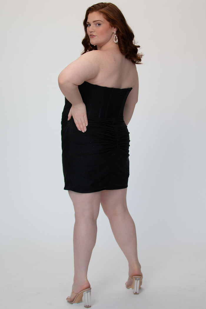 Short fitted black party dress SC8143 by Sydney's Closet. Back view.  Strapless, exposed boning basque waist and ruched fitted skirt in shimmer stretch knit. Availability in black, purple, red, ivory, green and royal. Model in picture is 5'9.