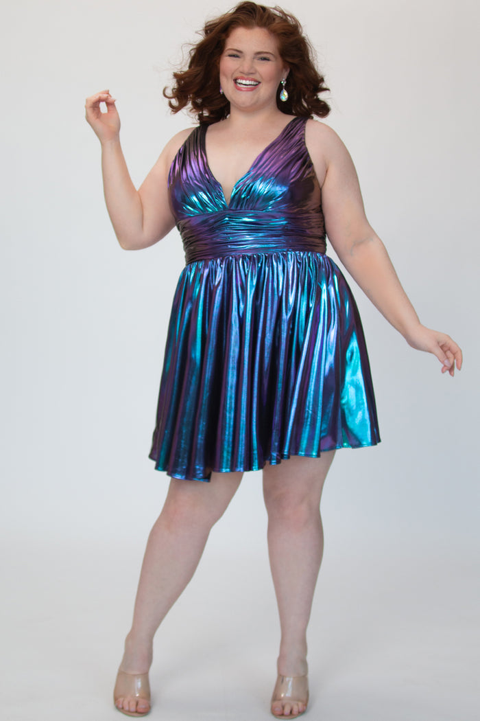 A-line flowy short party dress, SC8156 by Sydney's Closet. Front view. In the color Prism, a blue/purple knit metallic fabric. Thick bra-friendly straps, pleated v-neckline bodice and waist band, long invisible center back zipper. V- back. Available in Prism, Peacock and Raven black, sizes 14-32. 
Model in picture is 5'9. Model in picture is 5'9.