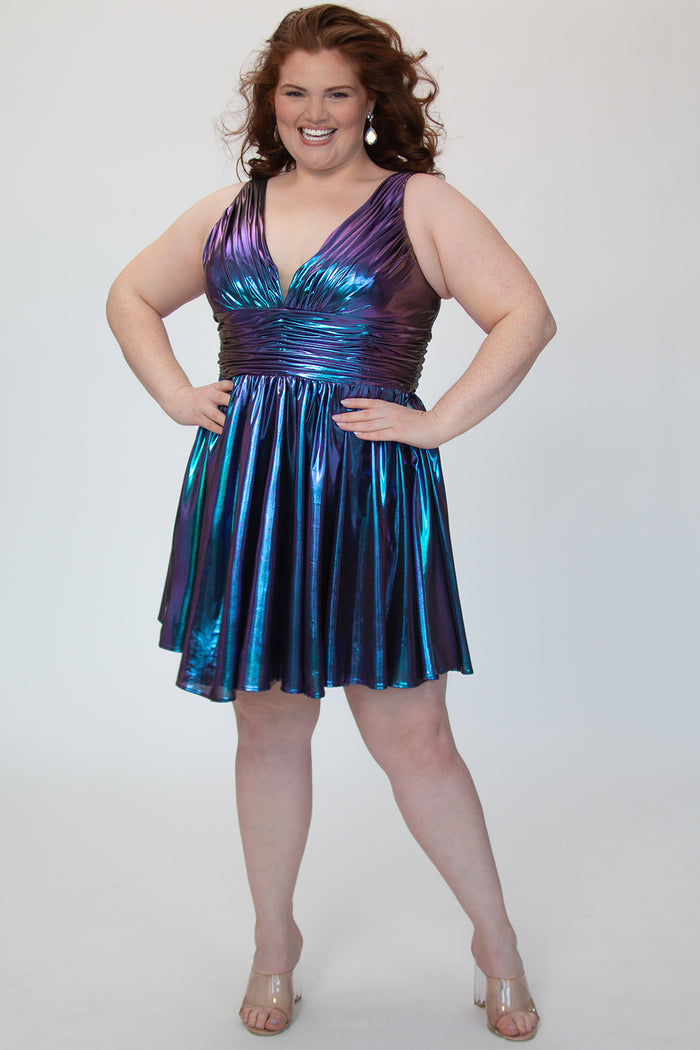 A-line flowy short party dress, SC8156 by Sydney's Closet. Front view. In the color Prism, a  blue/purple metallic knit fabric. Thick bra-friendly straps, pleated v-neckline bodice and waist band, long invisible center back zipper. V- back. Available in Prism, Peacock and Raven black, sizes 14-32. 
Model in picture is 5'9.