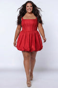 Plus-size model wearing a red sleeveless taffeta mini dress with a square neckline, structured corset-inspired bodice with exposed boning, and voluminous bubble hem skirt. She is styled with long flowing brown hair, drop earrings, and nude platform ankle-strap heels, walking toward the camera against a light gray studio background. Front view.

Sydney's Closet SC8166 in red