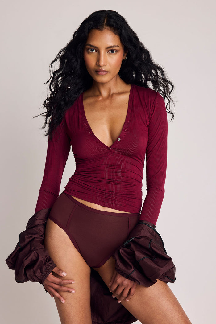 A model wearing a ribbed long-sleeve wrap-style bodysuit in deep burgundy with matching high-waisted briefs and dramatic ruffled sleeves, posed against a light gray studio background.