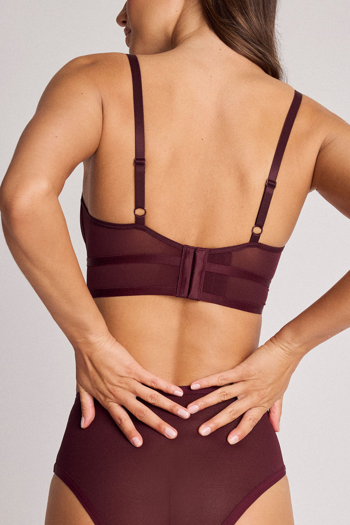 Rear view of a woman wearing a dark burgundy mesh and fabric bralette with adjustable shoulder straps and high-waisted matching underwear against a light gray studio background.