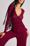 A woman wearing a ribbed long-sleeve top in deep burgundy, featuring a plunging V-neckline with small silver buttons, a gathered waist, and button details on the front of the pants, set against a solid light gray background.