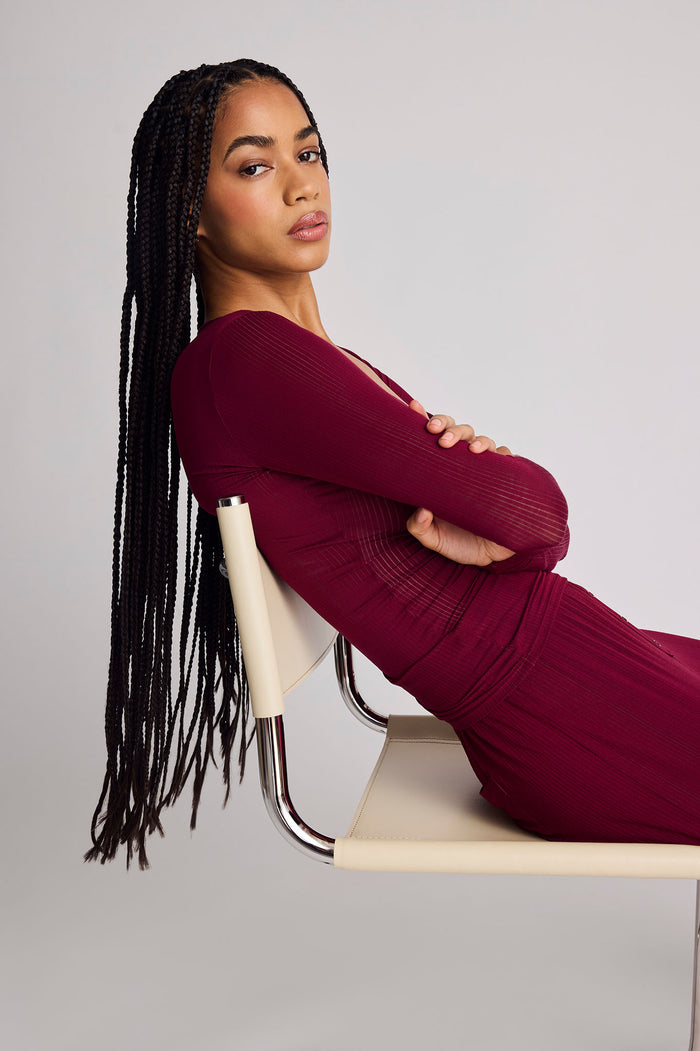 A woman with long black braids reclines on a cream-colored chair with a metallic frame, wearing a deep burgundy ribbed long-sleeve henley against a plain light gray studio background.